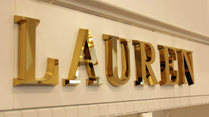 Gold metallic 3D letters spelling "LAUREN" mounted on a light-colored wall.